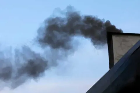 Gęsty, czarny dym wydobywa się z komina, tworząc kłęby na tle błękitnego nieba. Ten smród z komina w pokoju jest nie do zniesienia.