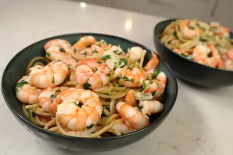 Pyszny makaron z krewetkami, posypany świeżą natką pietruszki i parmezanem, podany w ciemnych miseczkach.