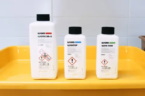 Three Ilford bottles for developing, stopping, and fixing film sit in a yellow tray, ready for creating the best black and white film.