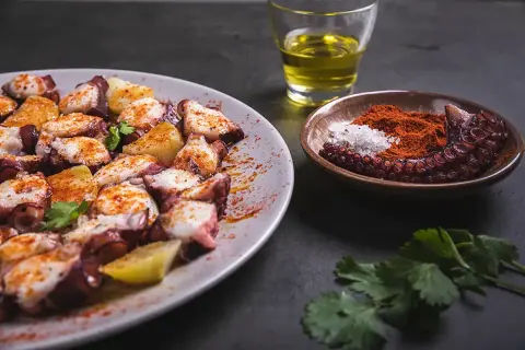 Un plat de poulpe à la gallega, garni de pommes de terre et d'épices, avec un petit bol de paprika et de sel, et un verre d'huile d'olive.