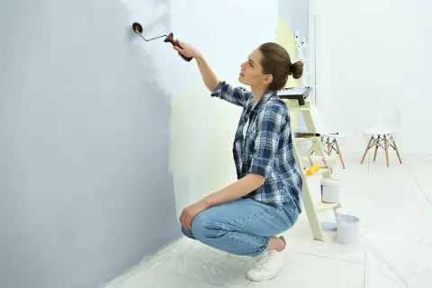 Młoda kobieta w kratę i jeansach maluje ścianę wałkiem. W tle drabina, puszki z farbą i krzesła.
