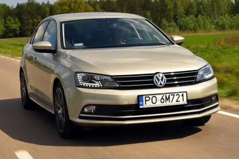 Złoty VW Jetta MK6 jedzie po drodze wśród zielonych pól i lasów.