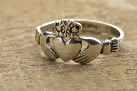 Anillo Claddagh de color oro blanco, con corazón, manos y corona, sobre madera.