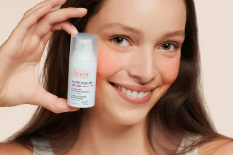 Ein lächelndes Gesicht hält eine Flasche Avène Antirougeurs. Ideal bei Rötungen, auch bei Neurodermitis.