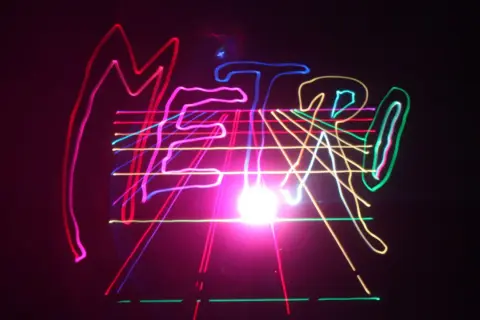Kolorowe, neonowe linie tworzą napis "METRO" na czarnym tle, przypominając o energetycznym, musical metro.