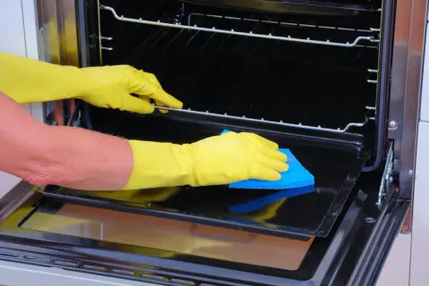 Manos con guantes amarillos limpiando el interior de un horno con una esponja azul.