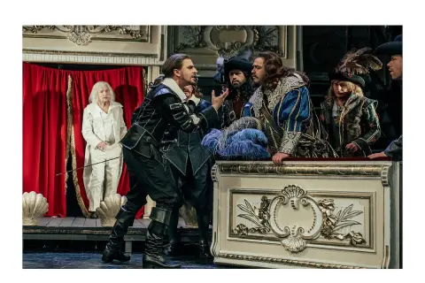 Aktorzy w strojach z epoki na scenie, w tym mężczyzna w czarnym stroju, przypominający Cyrano de Bergerac, gestykuluje do towarzyszy.