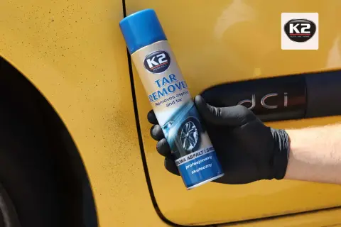 Rękawiczka z czarną gumą trzyma puszkę preparatu do dekontaminacji lakieru. Żółty lakier samochodu jest pokryty drobnymi, ciemnymi plamkami smoły.