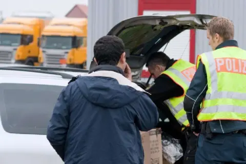 Polizisten durchsuchen ein Auto. Ein Mann telefoniert, während die Beamten den Kofferraum inspizieren. Im Hintergrund stehen Lastwagen.