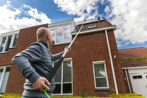 Hombre con pértiga extendida para limpiar tejas de una casa de ladrillo rojo bajo un cielo azul con nubes.