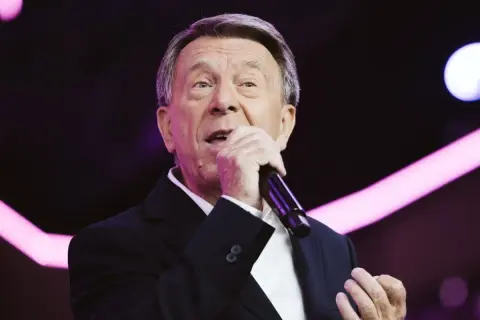 Wojciech Gąssowski śpiewa "Gdzie się podziały tamte prywatki". Artysta w czarnym garniturze i białej koszuli, z mikrofonem w dłoni, na tle fioletowych świateł.