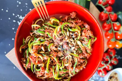 Ein leckeres Zoodles Rezept mit Hackfleischsauce, Pinienkernen und Parmesan in einer roten Schüssel. Daneben Kirschtomaten und Zucchini.