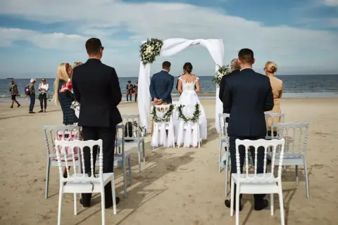 Piękny ślub na plaży. Para młoda stoi przed białym baldachimem, goście obserwują ceremonię.