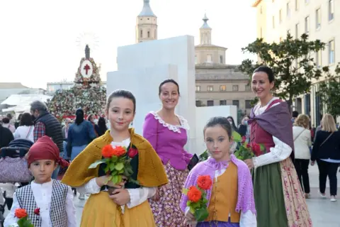 Niñas con trajes regionales y flores rojas, listas para las fiestas Zaragoza. Detrás, un gran tapiz floral y edificios históricos.