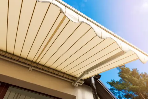 Toldo beige desplegado sobre terraza, cumpliendo la normativa. Cielo azul y árbol al fondo.