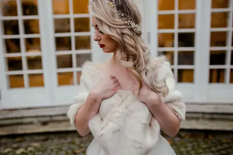 Elegancka panna młoda w białej etoli z futra, ozdobiona perłami i złotymi elementami we włosach, pozuje przed drzwiami.