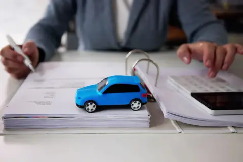 Osoba z długopisem i kalkulatorem analizuje dokumenty, obok stoi niebieski model auta. Zastanawia się, po jakim czasie można sprzedać auto.