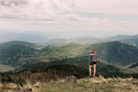 Una excursionista con sombrero admira las majestuosas montañas de Madrid, un paisaje verde y brumoso bajo un cielo nublado.