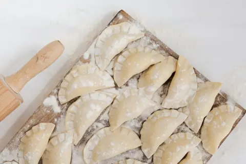 Surowe pierogi ruskie ułożone na desce, obok wałek do ciasta.