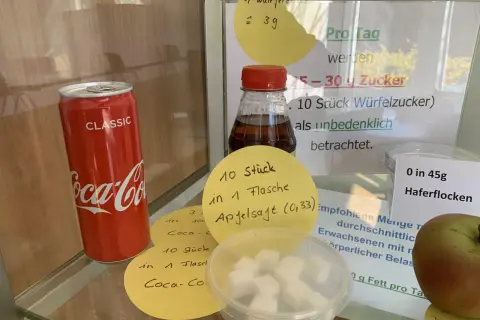 Eine Cola-Dose neben Würfelzucker. Die Frage "wie viel gramm zucker hat cola" wird durch Notizen beantwortet: 10 Stück Würfelzucker pro Flasche.