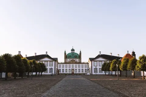 Frederiksborgs slott med sin gröna kupol reser sig majestätiskt mot en klar himmel. En stenlagd väg leder fram till den imponerande byggnaden, flankerad av träd.