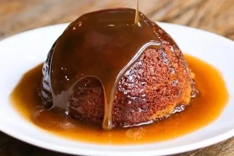 Un delizioso sticky toffee pudding, ricoperto da una cascata di salsa al caramello, servito su un piatto bianco.