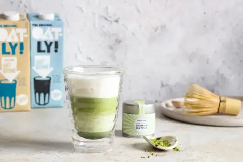 Warstwowy napój matcha z mlekiem owsianym, obok puszka z matchą i bambusowa miotełka. Idealny matcha przepis.