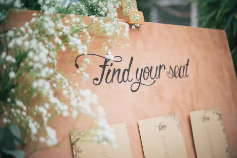 Panneau en bois avec l'écriture "Find your seat" et des fleurs. Des cartes de placement sont fixées en dessous.