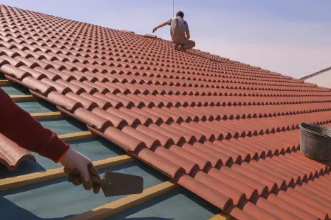 Un tejado en proceso de colocación de tejas rojas. Un trabajador con guantes usa una paleta, mientras otro se sienta en la cima.