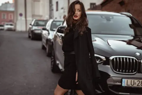 Elegancka kobieta w czarnej sukience swetrowej i płaszczu stoi obok luksusowego BMW na ulicy.