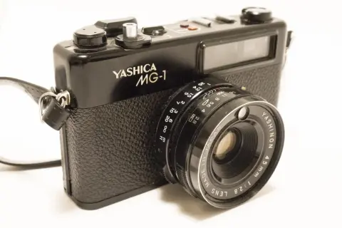 A black Yashica MG-1 camera with a leather strap. The lens reads "YASHINON 45mm 1:2.8".