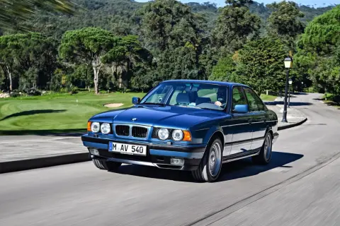 Niebieskie BMW E34 z lat 90. sunie po krętej drodze, mijając malownicze pole golfowe.