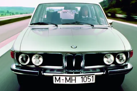 Klasyczne bmw e3 pędzi autostradą. Kierowca w garniturze pewnie trzyma kierownicę.