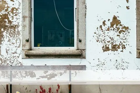 Paredes con desconchones y manchas de humedad, evidencia de filtraciones de agua de lluvia. Una ventana refleja el interior y un banco se sitúa delante.
