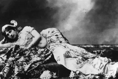 Mata Hari, bailarina exótica, yace recostada sobre telas estampadas, con un tocado adornado y mirada cautivadora.