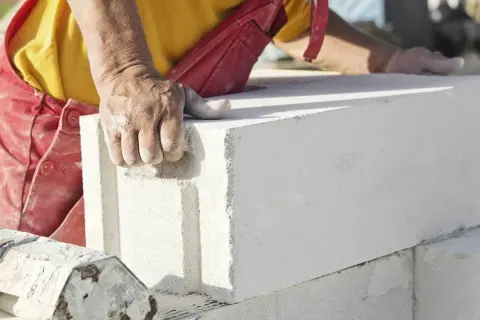 Dłoń pracownika w czerwonym fartuchu chwyta biały blok z betonu komórkowego, przygotowując go do budowy.