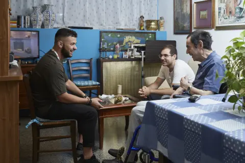 Tre uomini discutono in un salotto. Uno di loro, seduto su una sedia a rotelle, potrebbe chiedere quanto prende di pensione un dializzato.