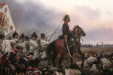 Soldados en formación, con un estandarte y un oficial a caballo, en medio de la **batalla de Bailén**. Cuerpos yacen en el suelo.