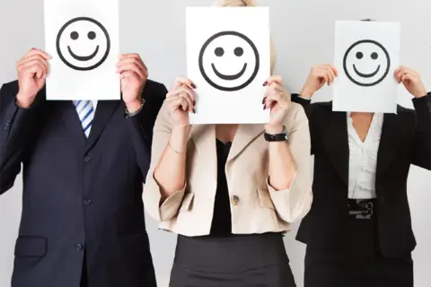 Trois personnes en tenue professionnelle cachent leur visage derrière des feuilles avec des smiley. Le bonheur est une idée neuve en Europe, et ces visages souriants le prouvent.