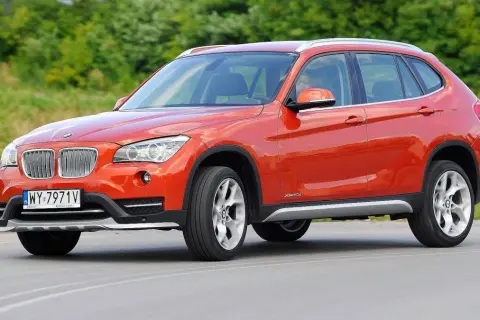 Pomarańczowe BMW E84 X1 z polskimi tablicami rejestracyjnymi jedzie po drodze, otoczone zielenią.