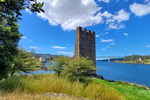 Torre medieval junto a un río azul en Catoira, Galicia. Un puente cruza el agua al fondo.
