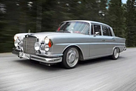 Silver Mercedes från 60-talet glider fram på en väg, omgiven av skog. Klassiska bilar som denna är tidlösa.