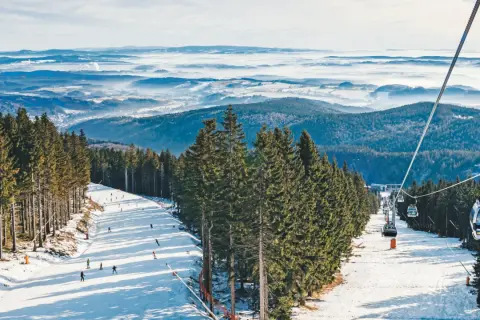 Skifahrer genießen den Winter in Tschechien. Eine Seilbahn schwebt über schneebedeckte Hänge, umgeben von dichten Fichtenwäldern und einer nebligen Berglandschaft.