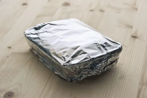 Ein rechteckiges Paket, in glänzende Alufolie gewickelt, liegt auf einem Holztisch. Die Frage, welche Seite der Alufolie giftig ist, bleibt unbeantwortet.