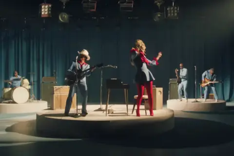 Lady Gaga w czerwonych rajstopach i niebieskiej sukience tańczy na scenie, podczas gdy zespół gra. Tekst "die with a smile" może być inspiracją do tej sceny.