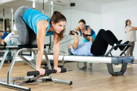 Frauen trainieren im Studio. Eine macht eine Übung auf einem Gerät, während andere im Hintergrund an Pilates-Geräten arbeiten. Das ist Zirkeltraining.