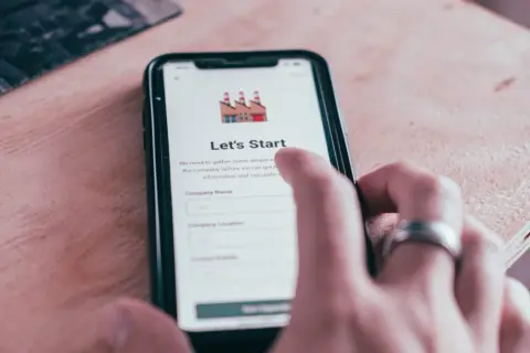 Dłoń z pierścionkiem dotyka ekranu smartfona z aplikacją "Let's Start". Aplikacja zbiera dane firmy, wysyłając powiadomienia.
