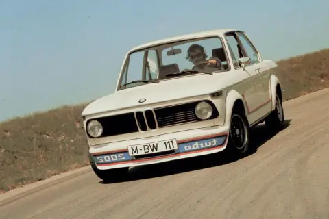 Białe BMW E20 z kolorowymi pasami na zderzaku i czerwonymi akcentami na karoserii jedzie po krętej drodze.