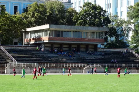 Młodzi piłkarze trenują na boisku. W tle trybuny i budynek, przypominający stadion Dynamo Kijów.