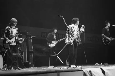 Vier Musiker auf einer Bühne, die alte Rockbands repräsentieren. Einer spielt Schlagzeug, die anderen Gitarre und Bass.